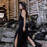 A woman in the AYA Sacred Wear Black Cotton Bohemian Dress, paired with a black hat and boots, walks outdoors on stone steps beside a rustic bamboo structure.