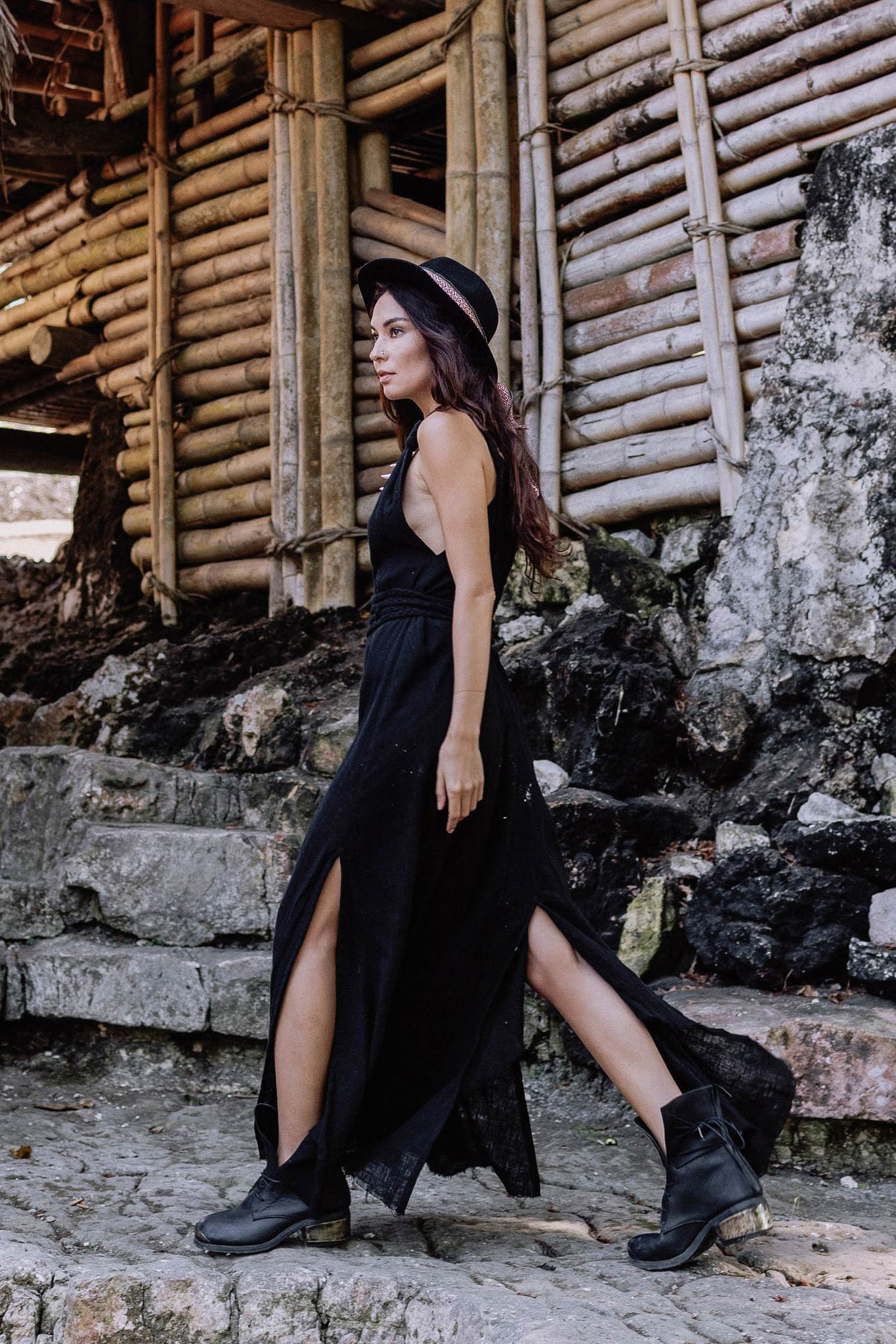 A woman in the AYA Sacred Wear Black Cotton Bohemian Dress, paired with a black hat and boots, walks outdoors on stone steps beside a rustic bamboo structure.