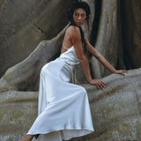 A woman in a flowing White Peace Silk Simple Wedding Dress by AYA Sacred Wear poses gracefully against a large tree trunk with textured bark. Her dark hair is styled in a braid, and she leans forward with one foot on the ground, conveying elegant strength, like a vision from a bohemian wedding dream.