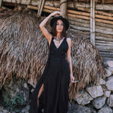 A woman stands confidently outdoors by a rustic bamboo structure, wearing the Black Cotton Bohemian Dress by AYA Sacred Wear. She pairs it with a statement necklace and hat, embodying effortless style in organic handwoven cotton.