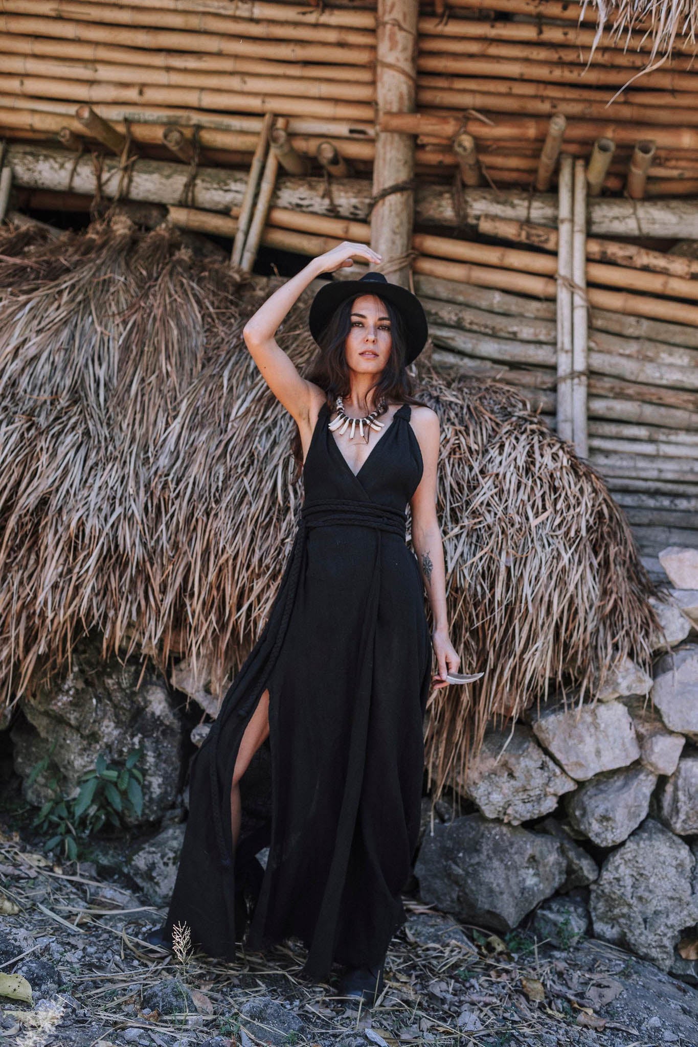 A woman stands confidently outdoors by a rustic bamboo structure, wearing the Black Cotton Bohemian Dress by AYA Sacred Wear. She pairs it with a statement necklace and hat, embodying effortless style in organic handwoven cotton.