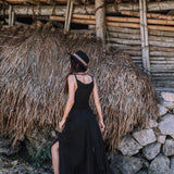 A woman wearing the AYA Sacred Wear Black Cotton Bohemian Dress and a hat stands with her back to the camera before a rustic bamboo, stone, and dried thatch structure.
