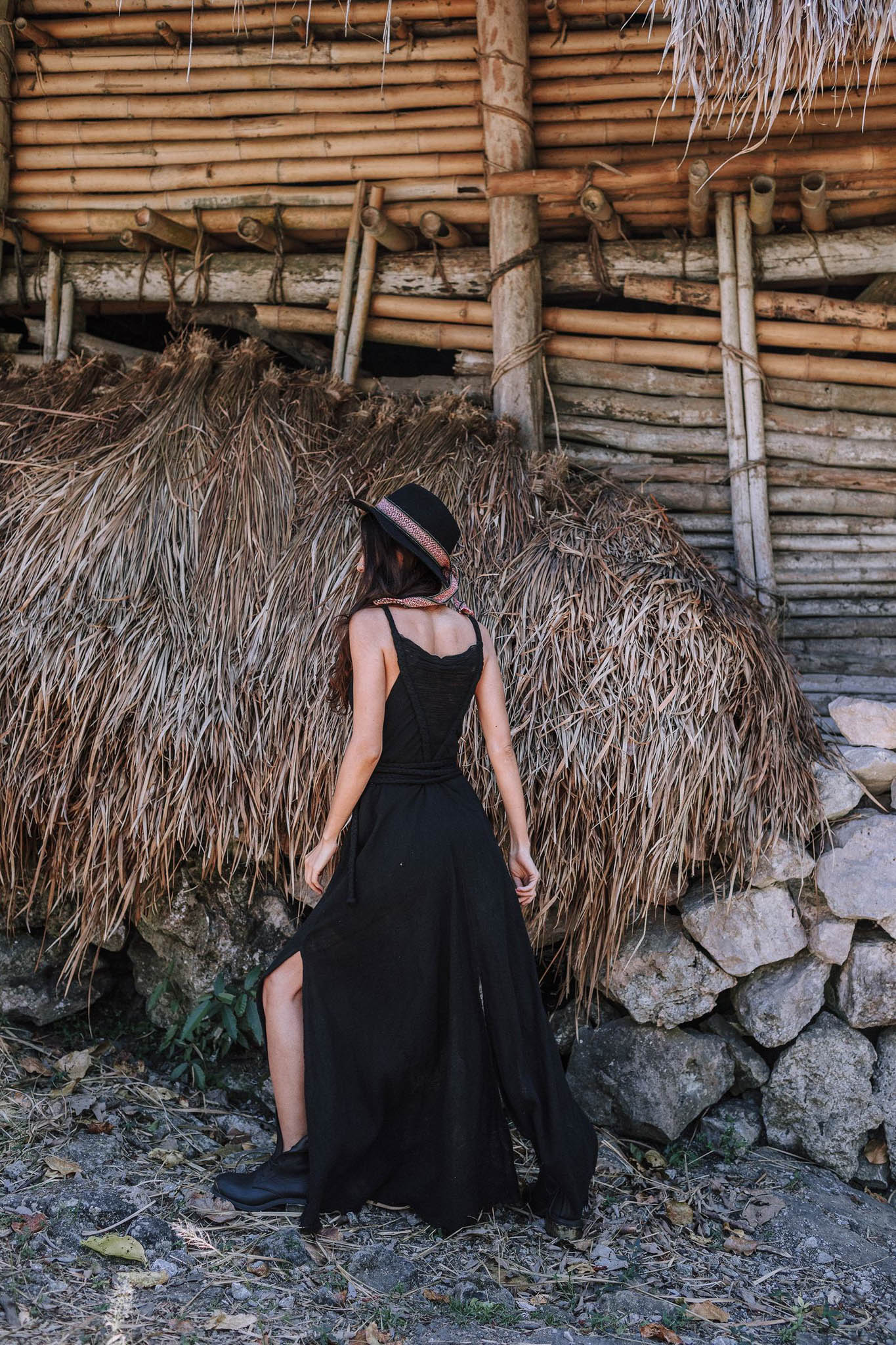A woman wearing the AYA Sacred Wear Black Cotton Bohemian Dress and a hat stands with her back to the camera before a rustic bamboo, stone, and dried thatch structure.