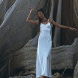 A woman in a flowing, bohemian dress from AYA Sacred Wear's White Peace Silk Simple Wedding Dress collection stands barefoot on large tree roots. She is surrounded by the massive trunk of an ancient tree, with her arm raised gracefully and her long hair cascading down her back, creating a serene and natural scene.