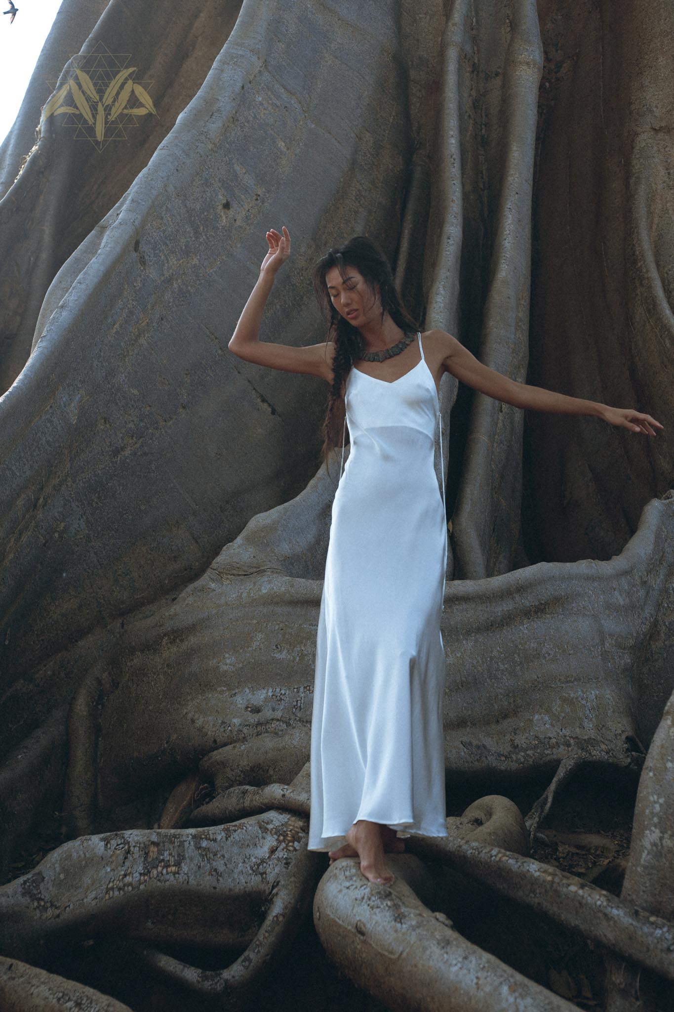 A woman in a flowing, bohemian dress from AYA Sacred Wear's White Peace Silk Simple Wedding Dress collection stands barefoot on large tree roots. She is surrounded by the massive trunk of an ancient tree, with her arm raised gracefully and her long hair cascading down her back, creating a serene and natural scene.