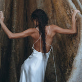 A woman wearing the White Peace Silk Simple Wedding Dress by AYA Sacred Wear stands between two large, smooth rocks. Her long hair is styled in a braid that falls down her back as she faces away, touching the rocks with both hands. The scene is serene and natural.