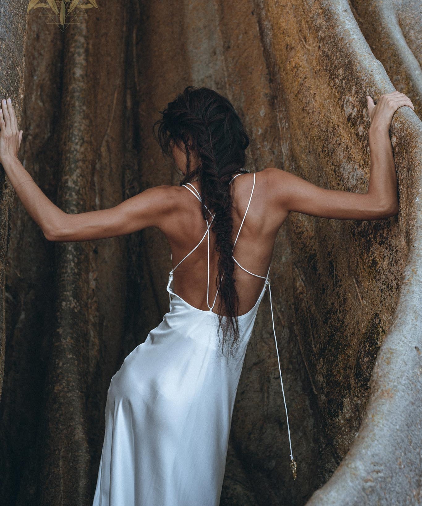 A woman wearing the White Peace Silk Simple Wedding Dress by AYA Sacred Wear stands between two large, smooth rocks. Her long hair is styled in a braid that falls down her back as she faces away, touching the rocks with both hands. The scene is serene and natural.
