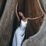 A woman wearing the White Peace Silk Simple Wedding Dress by AYA Sacred Wear leans against the large roots of a tree. The textures of the roots contrast beautifully with the smoothness of her natural silk boho dress. She gracefully poses with one arm elegantly raised above her head, embodying a seamless blend of nature and elegance.