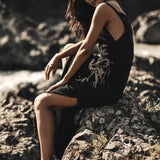 A woman in the AYA Sacred Wear Black Cotton Bohemian Dress sits barefoot on rocky terrain among wild plants, sunlight casting shadows that highlight her relaxed, contemplative pose.