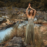 A woman in the Sage Green Tribal Goddess Slip Over by AYA Sacred Wear stands on rocks beside a stream, surrounded by lush greenery and boulders, her arms gracefully raised overhead.