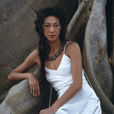 Wearing a White Peace Silk Simple Wedding Dress from AYA Sacred Wear, the woman with long, dark hair sits gracefully against a large, textured rock formation. The natural silk boho dress complements her chunky necklace, enhancing the earthy ambiance of the setting.