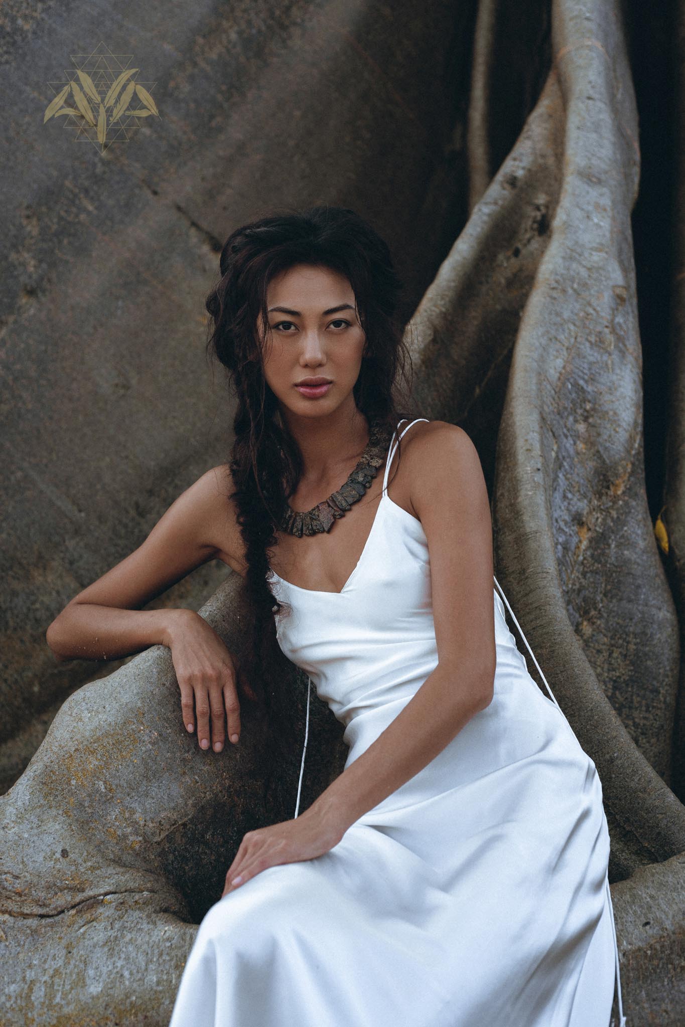 Wearing a White Peace Silk Simple Wedding Dress from AYA Sacred Wear, the woman with long, dark hair sits gracefully against a large, textured rock formation. The natural silk boho dress complements her chunky necklace, enhancing the earthy ambiance of the setting.