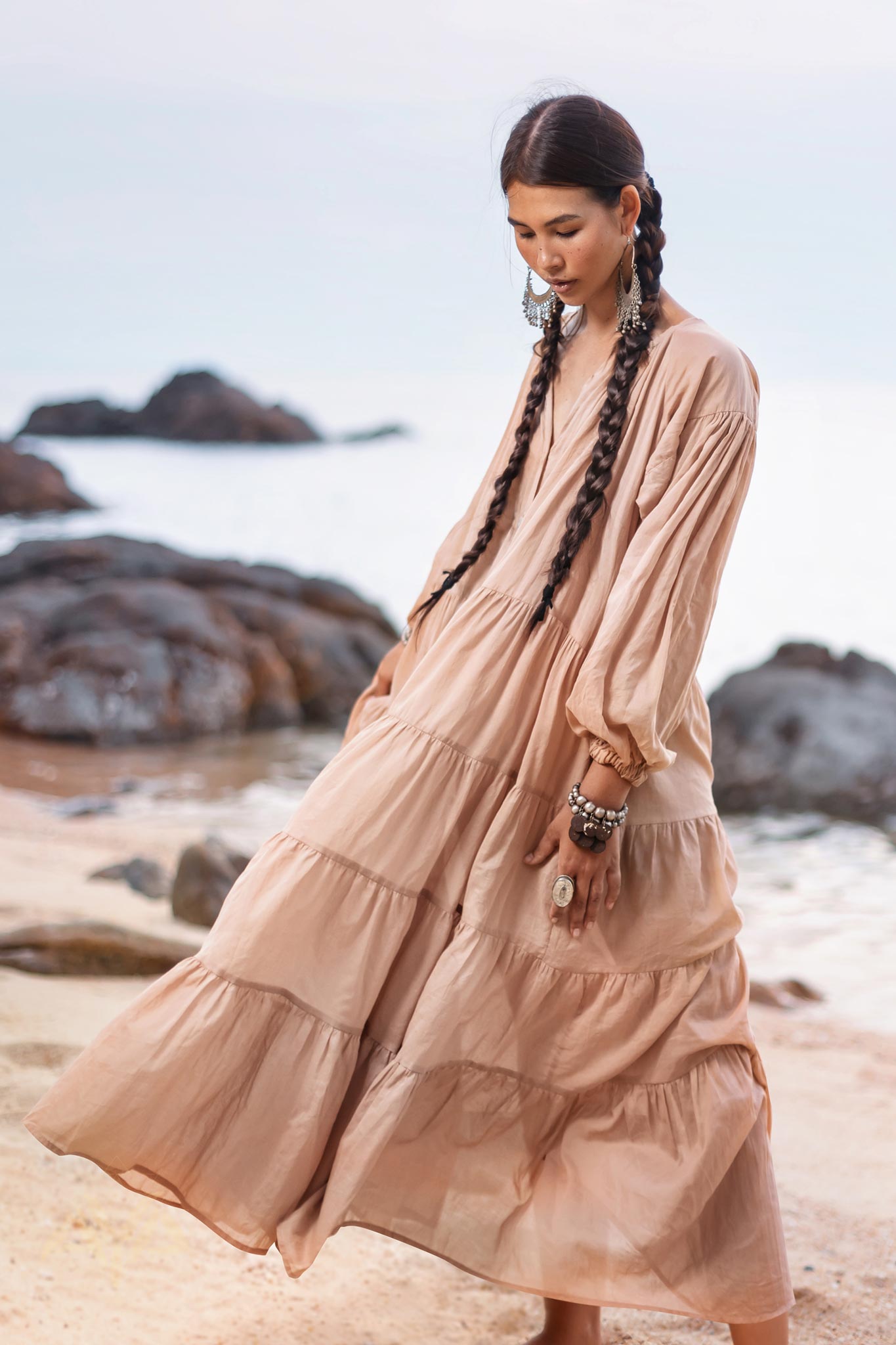 A woman with long braided hair wears an AYA Sacred Wear Boho Dress for Women in dusty pink. She stands thoughtfully on a rocky beach. Large earrings and bracelets complement her look, with the sea and a cloudy sky visible in the distance.