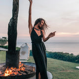 A barefoot woman dances joyfully at sunset by a fire pit, wearing the Black Cotton Bohemian Dress from AYA Sacred Wear, with the ocean and greenery in the background.