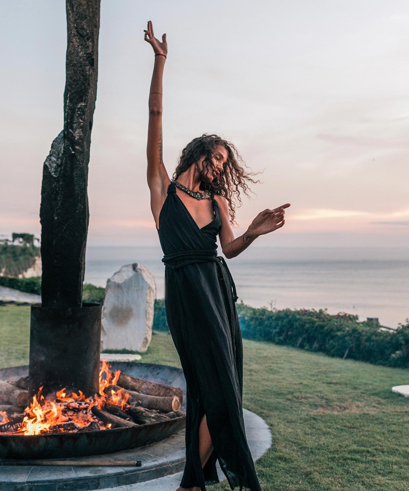 A barefoot woman dances joyfully at sunset by a fire pit, wearing the Black Cotton Bohemian Dress from AYA Sacred Wear, with the ocean and greenery in the background.