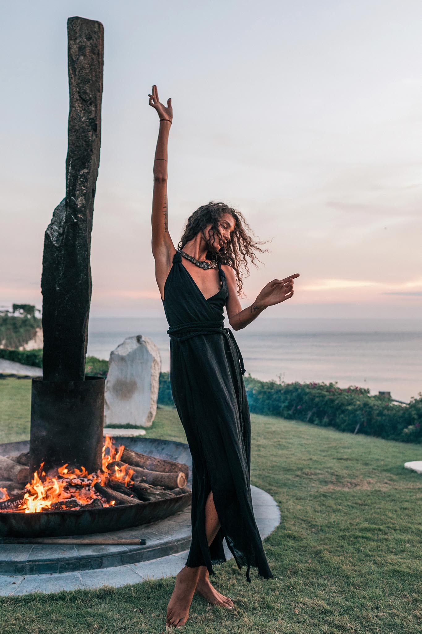 A barefoot woman dances joyfully at sunset by a fire pit, wearing the Black Cotton Bohemian Dress from AYA Sacred Wear, with the ocean and greenery in the background.