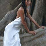 Wearing the White Peace Silk Simple Wedding Dress by AYA Sacred Wear, a woman poses against a textured stone backdrop. Her long braided hair elegantly drapes over her shoulder as she leans slightly to one side, with one hand gently resting on the stone surface, creating a natural and rugged setting.