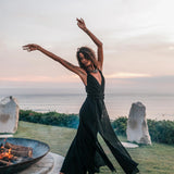 A woman wearing the AYA Sacred Wear Black Cotton Bohemian Dress dances barefoot on grass by a fire pit at sunset, with the ocean, pastel sky, and two large stones in the background.