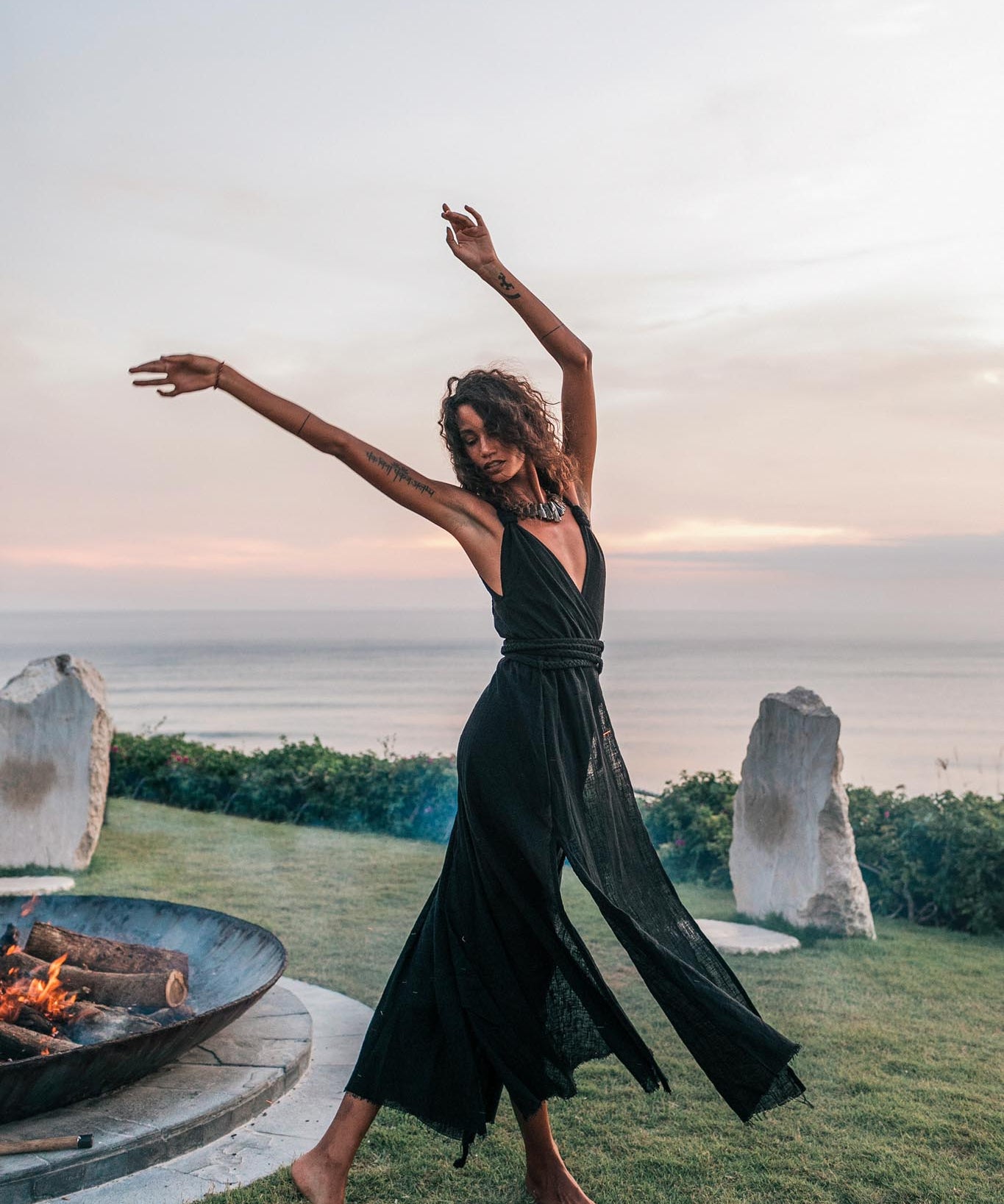 A woman wearing the AYA Sacred Wear Black Cotton Bohemian Dress dances barefoot on grass by a fire pit at sunset, with the ocean, pastel sky, and two large stones in the background.
