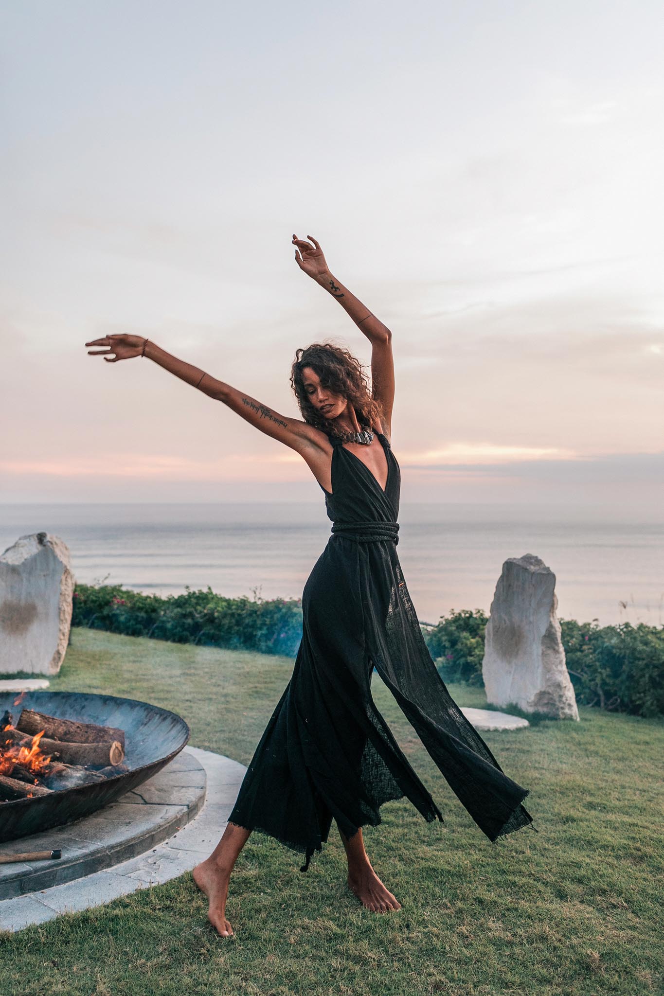 A woman wearing the AYA Sacred Wear Black Cotton Bohemian Dress dances barefoot on grass by a fire pit at sunset, with the ocean, pastel sky, and two large stones in the background.