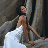 A woman wearing a White Peace Silk Simple Wedding Dress from AYA Sacred Wear leans back against a large, textured rock. Her eyes are closed, and her expression is serene. The flowing natural silk fabric contrasts with the rugged surface behind her.