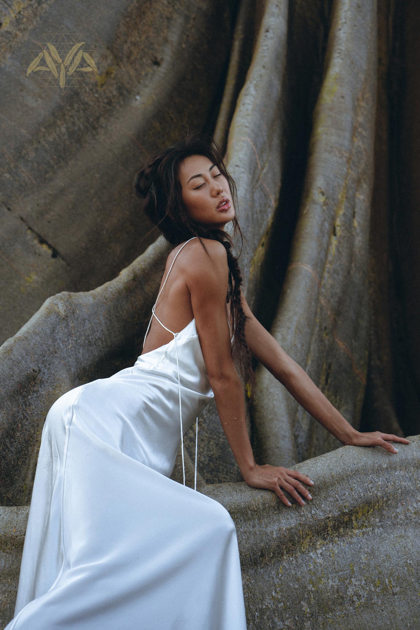 A woman wearing a White Peace Silk Simple Wedding Dress from AYA Sacred Wear leans back against a large, textured rock. Her eyes are closed, and her expression is serene. The flowing natural silk fabric contrasts with the rugged surface behind her.