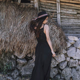 A woman in an AYA Sacred Wear Black Cotton Bohemian Dress and woven hat stands with her back turned before a rustic wood-and-thatch structure with a stone base.
