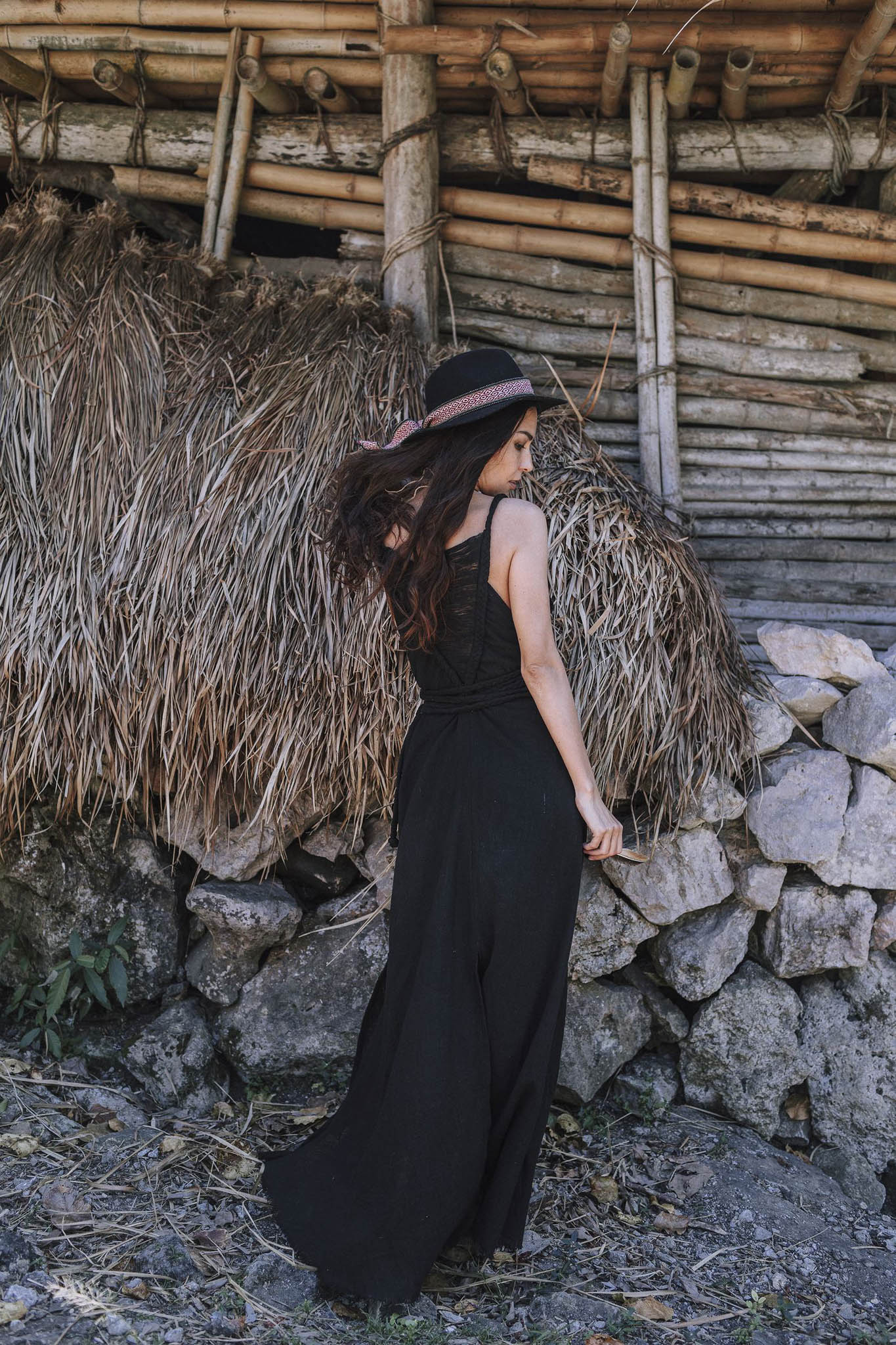 A woman in an AYA Sacred Wear Black Cotton Bohemian Dress and woven hat stands with her back turned before a rustic wood-and-thatch structure with a stone base.