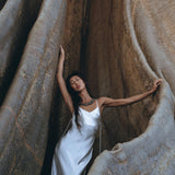 A woman wearing the elegant White Peace Silk Simple Wedding Dress by AYA Sacred Wear stands gracefully amid large rock formations. Her dark hair cascades over her shoulders as she gazes upward, enveloped by the textured, earthy tones of the rocks.