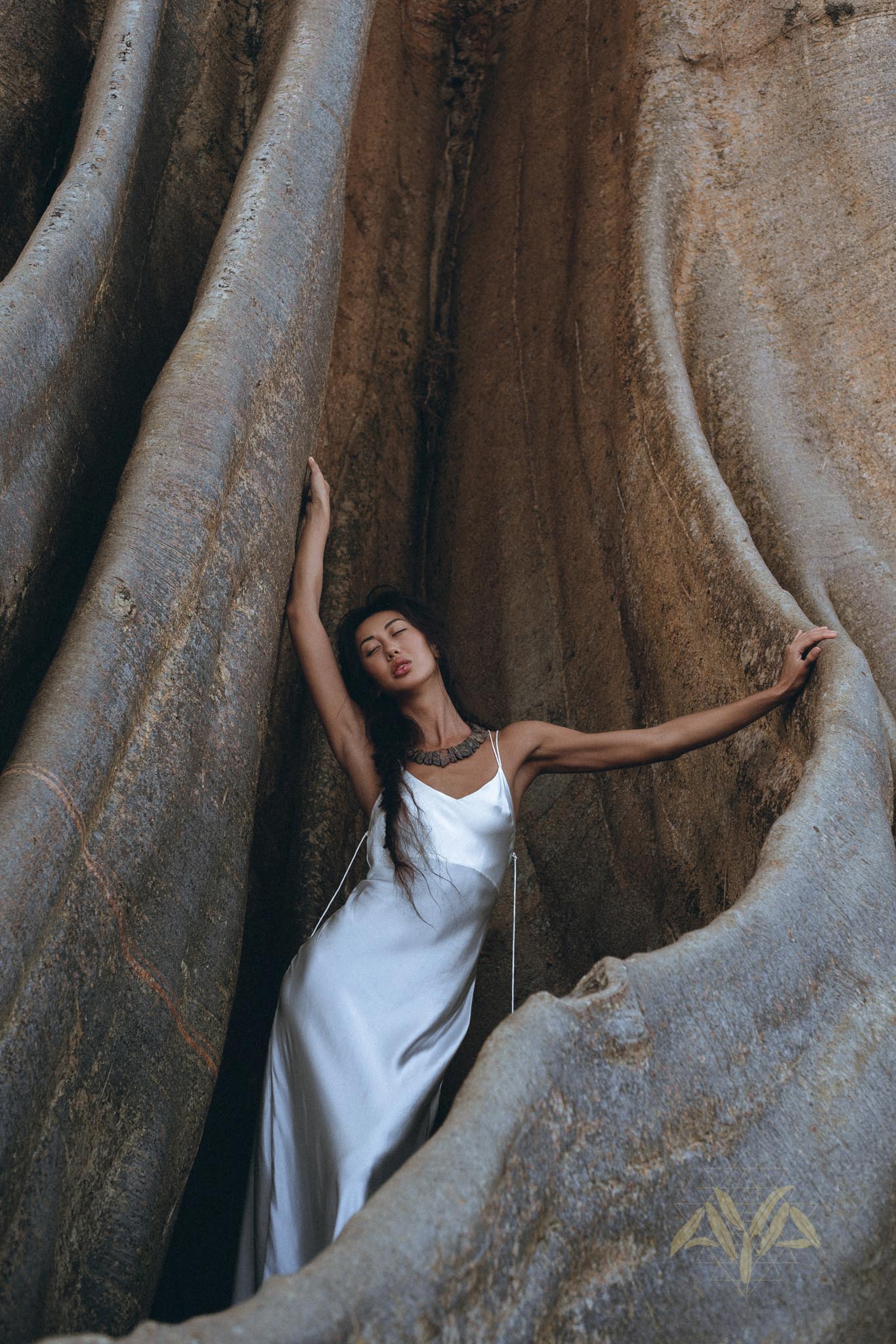 A woman wearing the elegant White Peace Silk Simple Wedding Dress by AYA Sacred Wear stands gracefully amid large rock formations. Her dark hair cascades over her shoulders as she gazes upward, enveloped by the textured, earthy tones of the rocks.