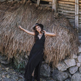 A woman in the Black Cotton Bohemian Dress by AYA Sacred Wear smiles, leaning against dried grass and a rustic bamboo-stone backdrop. She poses gracefully, one arm raised and the other outstretched in her bohemian wedding dress.