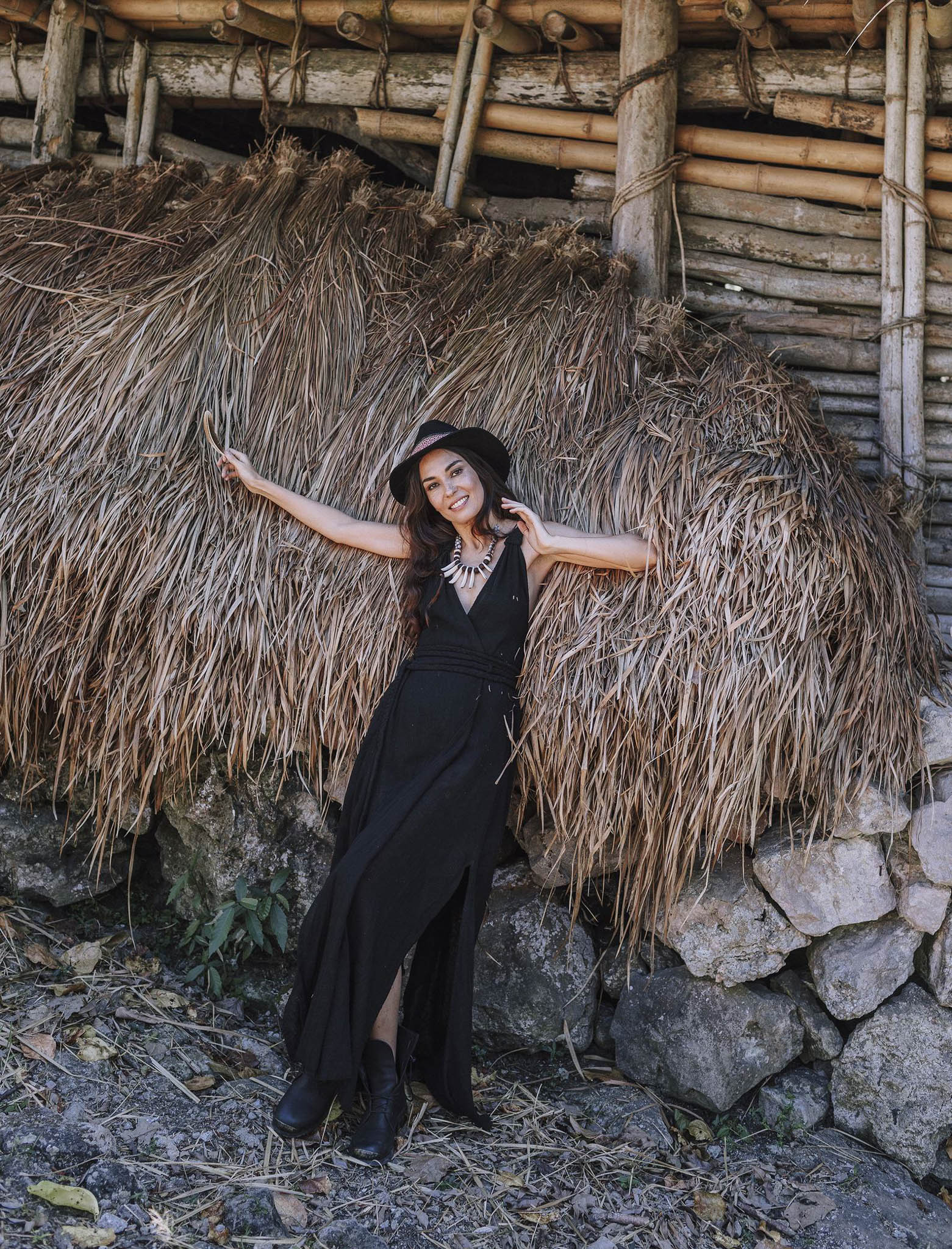 A woman in the Black Cotton Bohemian Dress by AYA Sacred Wear smiles, leaning against dried grass and a rustic bamboo-stone backdrop. She poses gracefully, one arm raised and the other outstretched in her bohemian wedding dress.