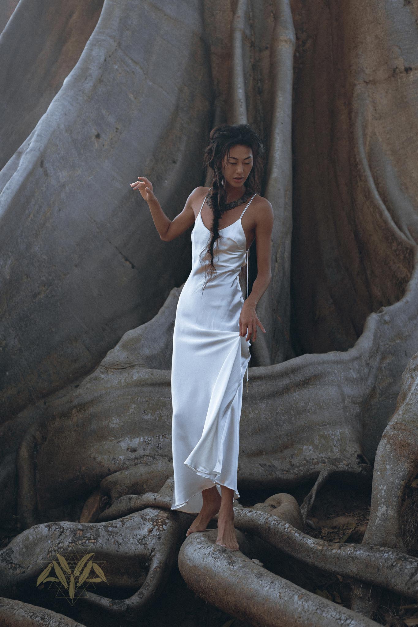 Draped elegantly in a white Peace Silk simple wedding dress from AYA Sacred Wear, a woman stands gracefully on the expansive roots of a gigantic tree. Her boho dress, with its natural silk fabric, harmonizes beautifully with the textures of the bark and roots surrounding her. The scene exudes a tranquil and earthy ambiance.