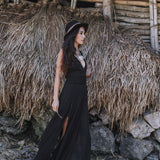 A woman in the Black Cotton Bohemian Dress by AYA Sacred Wear and a hat stands outdoors beside a rustic wooden and dried grass structure, with rocks and leaves scattered on the ground.