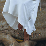 A person wearing the White Peace Silk Simple Wedding Dress by AYA Sacred Wear stands barefoot on large tree roots. The natural silk of the boho dress cascades gracefully, contrasting with the rugged texture of the roots.