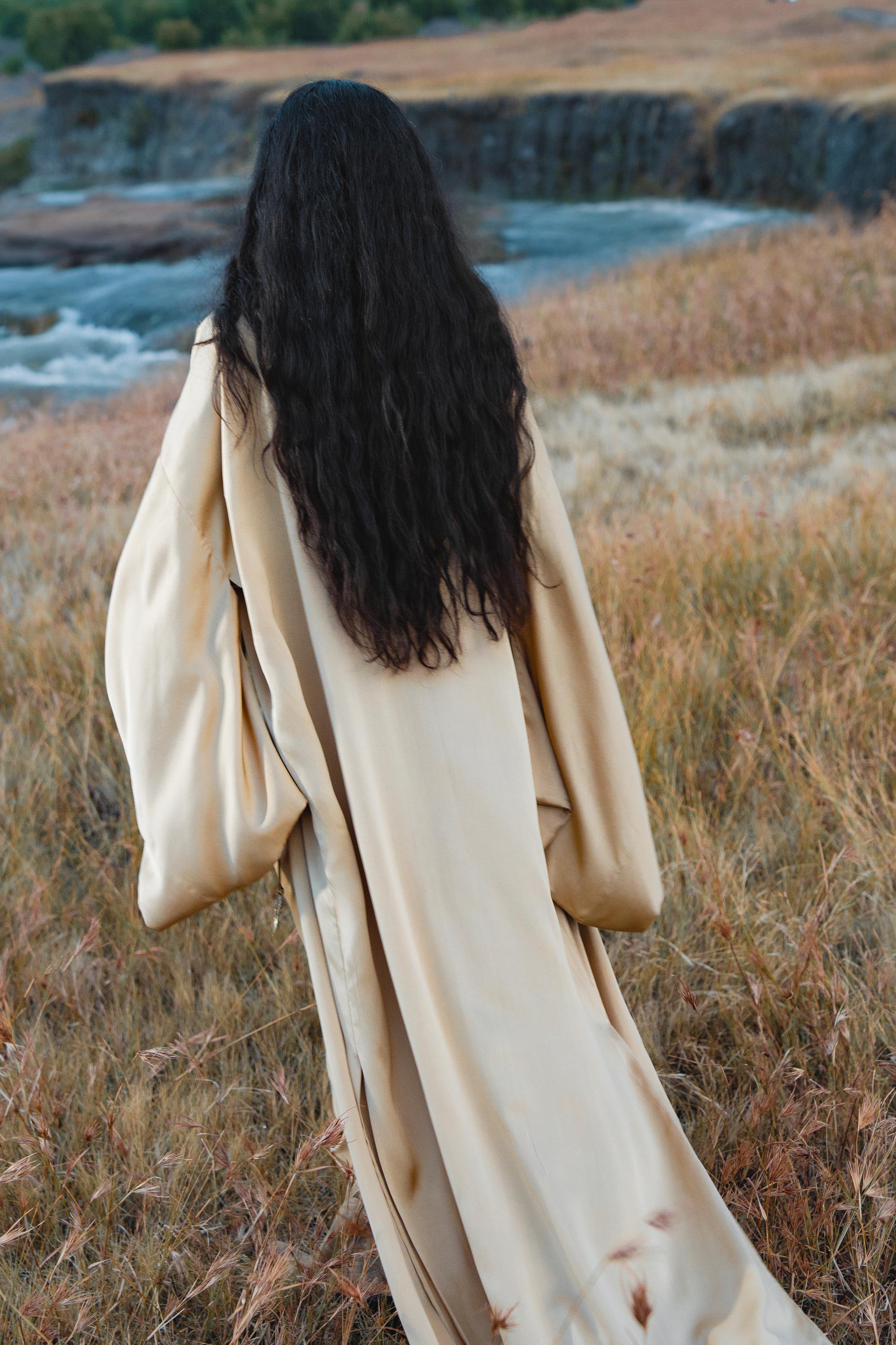 A person with long, wavy dark hair models the AYA Sacred Wear Gold Silk Long Robe—a floor-length, relaxed bohemian kimono—while standing in a dry, grassy field near a river, facing away from the camera.