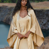 A woman with long dark hair stands outdoors by water, wearing the Gold Silk Long Robe—a floor-length maxi kimono from AYA Sacred Wear. She gazes down, hands clasped, amid grassy terrain and trees.
