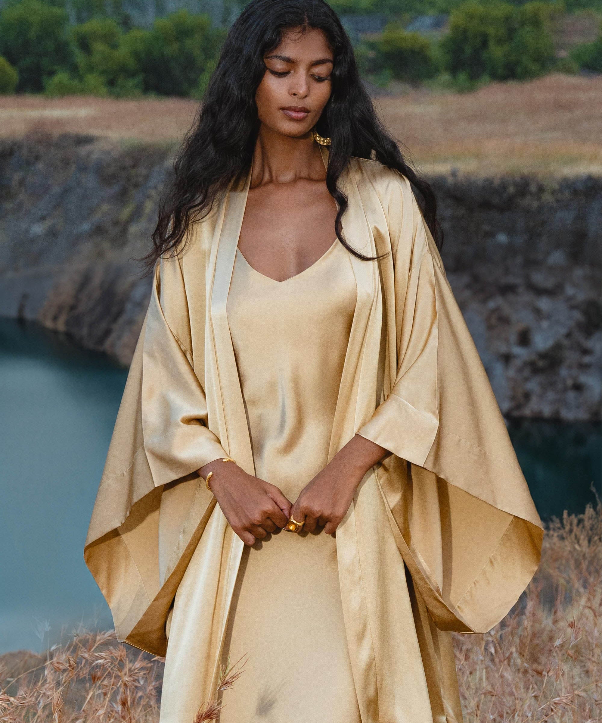 A woman with long dark hair stands outdoors by water, wearing the Gold Silk Long Robe—a floor-length maxi kimono from AYA Sacred Wear. She gazes down, hands clasped, amid grassy terrain and trees.