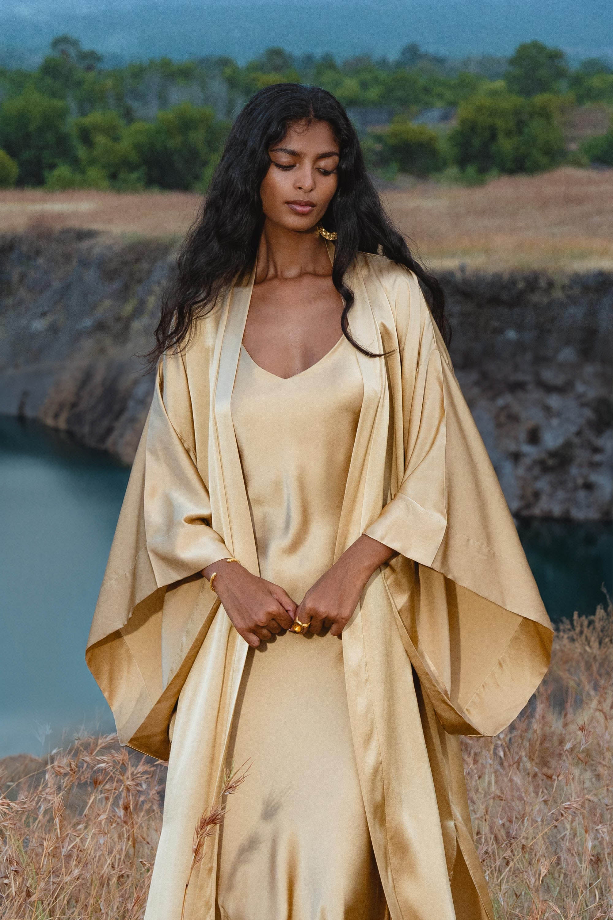 A woman with long dark hair stands outdoors by water, wearing the Gold Silk Long Robe—a floor-length maxi kimono from AYA Sacred Wear. She gazes down, hands clasped, amid grassy terrain and trees.
