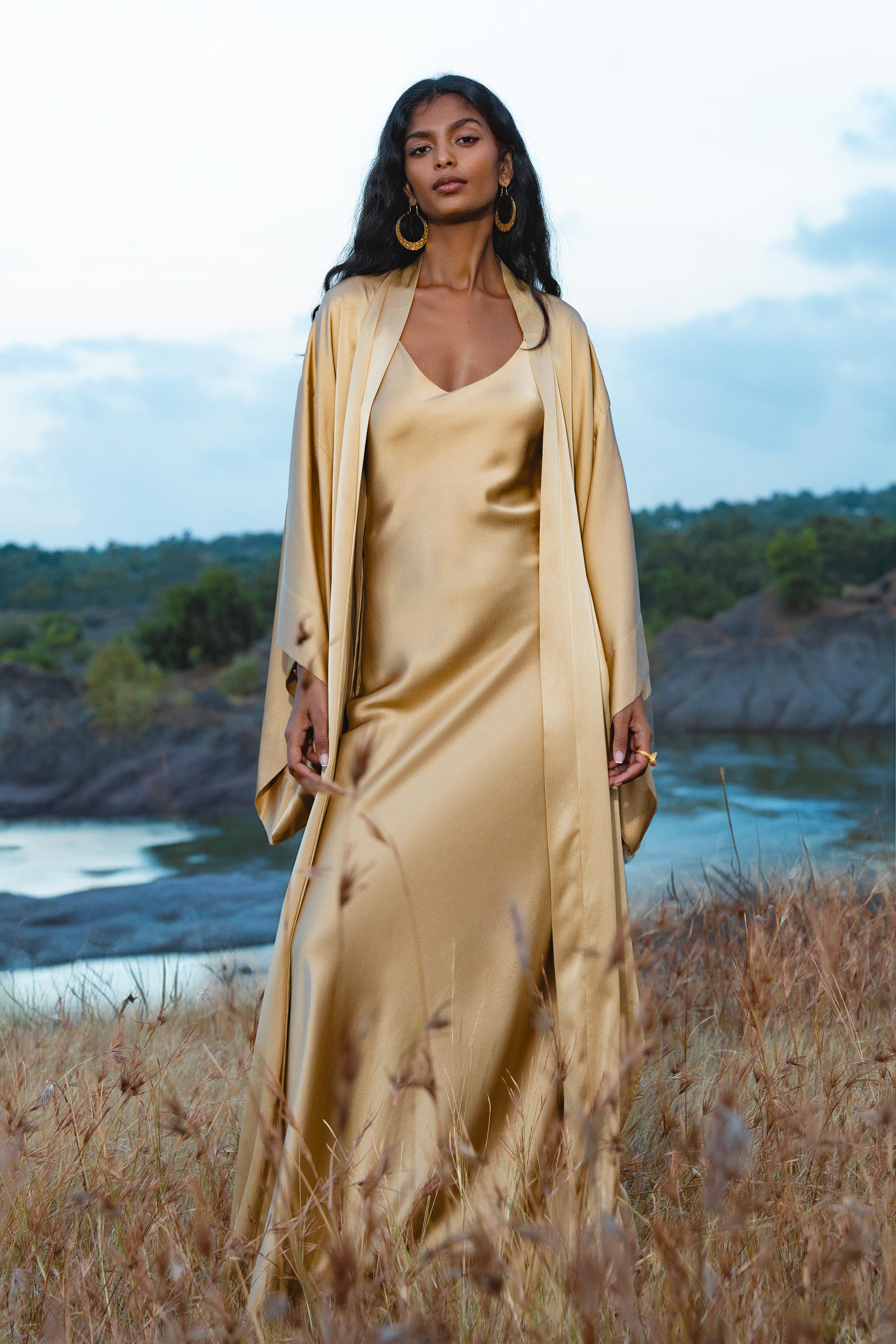 A woman with long dark hair and gold hoop earrings stands outdoors in tall grass, wearing the Gold Silk Long Robe— a floor-length maxi kimono by AYA Sacred Wear—against a serene backdrop of water, hills, and a cloudy sky.