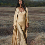 At sunset, a woman stands in a dry grassy field wearing the AYA Sacred Wear Gold Silk Long Robe — a floor-length maxi kimono and relaxed bohemian cardigan. Mountains and trees are visible beneath the partly cloudy sky in the background.