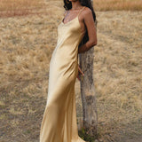 A woman in an AYA Sacred Wear Gold Silk Maxi Dress with a deep V-neck and backless design stands by a tree in a dry, grassy field with hills and trees in the background.