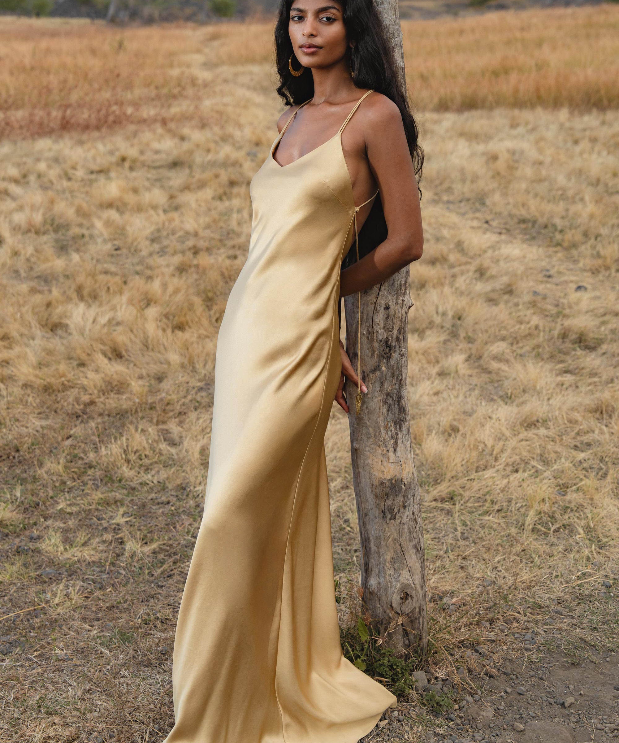 A woman in an AYA Sacred Wear Gold Silk Maxi Dress with a deep V-neck and backless design stands by a tree in a dry, grassy field with hills and trees in the background.