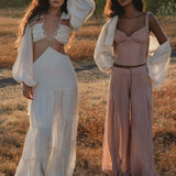Two women stand on dry grass in boho style—one wearing the AYA Sacred Wear Samudra Dress in Ivory Colour, an organic bohemian dress with long sleeves and cutouts; the other wears a mauve bustier, wide pants, and a sheer cover-up. Both have wavy hair with brass spiral accents.
