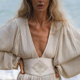 A woman with long, wavy blonde hair stands by the water in the Tatva Dress Ivory Colour by AYA Sacred Wear—a sustainable bohemian dress featuring voluminous sleeves, a deep V neckline, and an embroidered diamond-pattern belt.