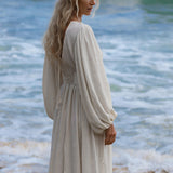 A woman with long, wavy blonde hair stands on the shore in the Tatva Dress Ivory Colour by AYA Sacred Wear, a sustainable bohemian cotton-linen dress with billowing sleeves, looking over her shoulder as gentle waves roll in behind her.