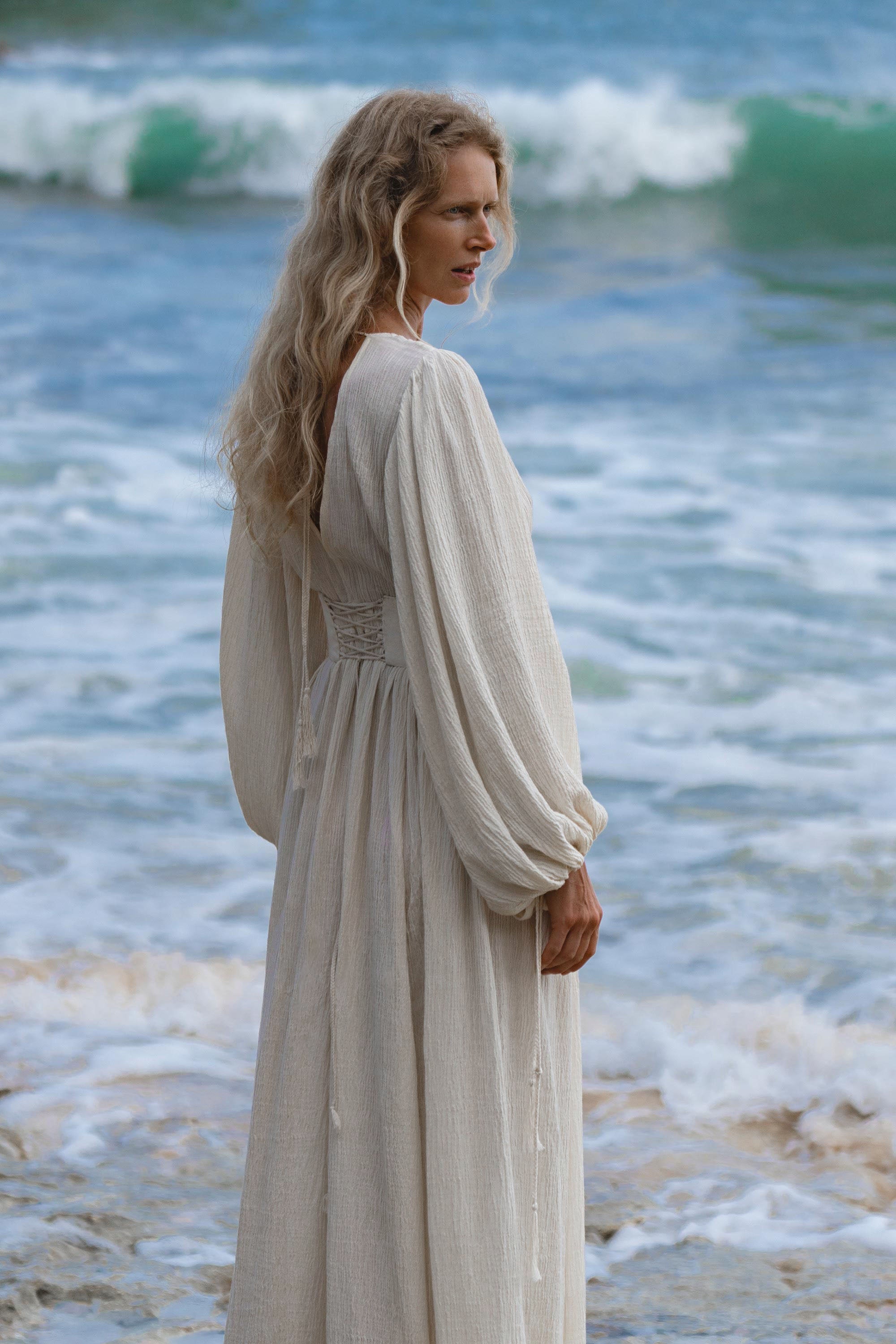 A woman with long, wavy blonde hair stands on the shore in the Tatva Dress Ivory Colour by AYA Sacred Wear, a sustainable bohemian cotton-linen dress with billowing sleeves, looking over her shoulder as gentle waves roll in behind her.