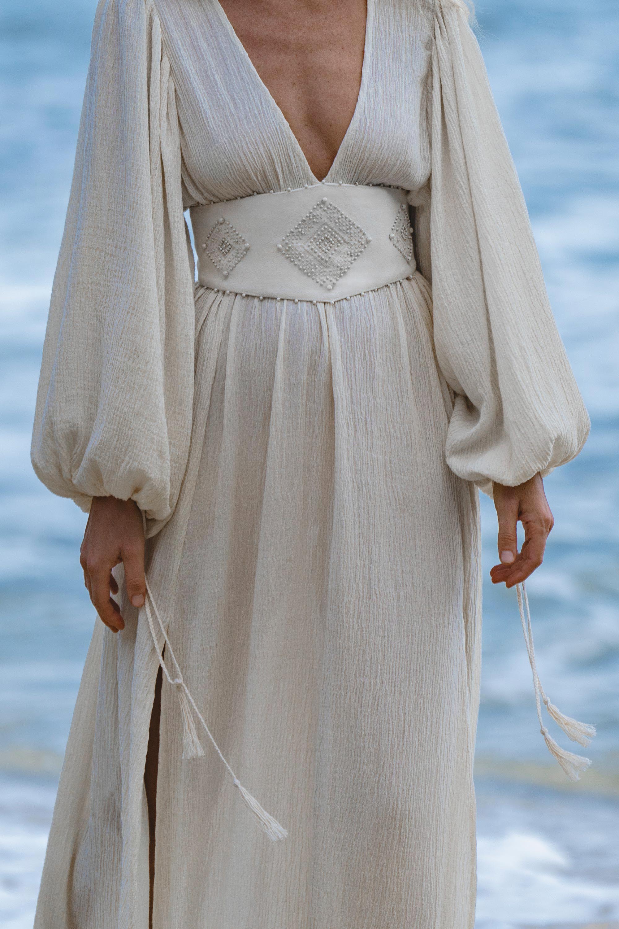 A person in the AYA Sacred Wear Tatva Dress Ivory Colour—a sustainable bohemian dress with natural cotton linen, billowy sleeves, deep V-neck, wide embroidered waistband, and side slit—stands by the blurred seaside with relaxed hands.