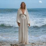 A person with long blonde hair stands on rocky sand near the ocean, wearing the Tatva Dress Ivory Colour—Sustainable Bohemian Dress by AYA Sacred Wear. Waves crash behind them as they gaze calmly at the camera under a cloudy blue sky.