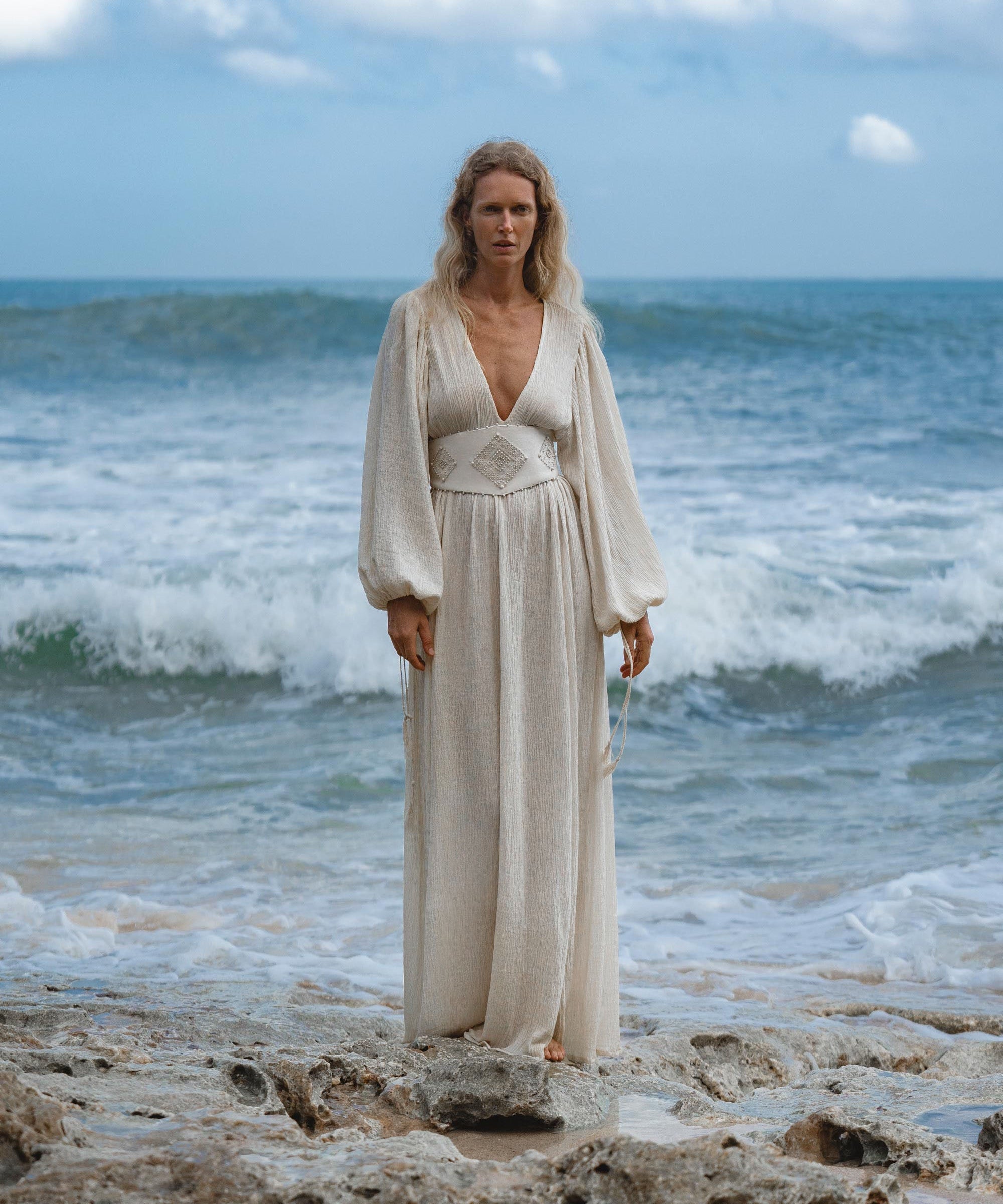 A person with long blonde hair stands on rocky sand near the ocean, wearing the Tatva Dress Ivory Colour—Sustainable Bohemian Dress by AYA Sacred Wear. Waves crash behind them as they gaze calmly at the camera under a cloudy blue sky.